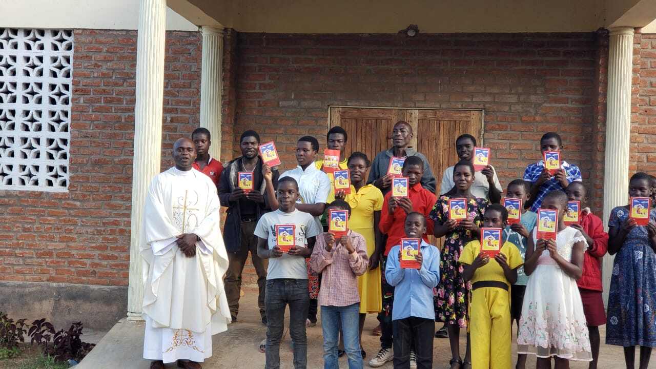 A group of people, including a person in white religious attire, stand in front of a building holding books.