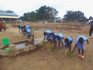 A group of children in uniforms are washing their hands at an outdoor water pump station in a rural setting.