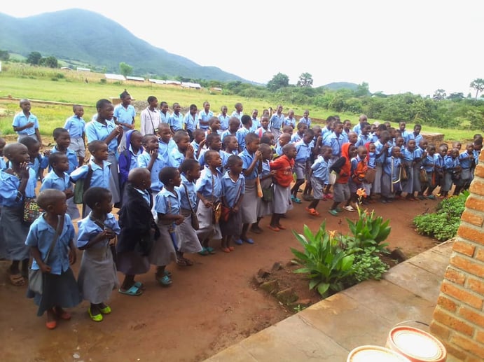 A large group of school children in uniforms stand in line outdoors, with a grassy area and hills in the background.