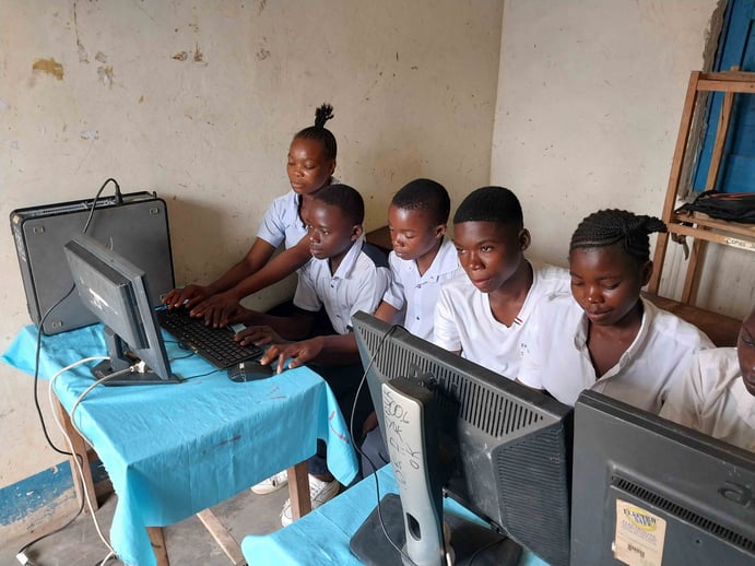 Five students in uniform sit closely together, using desktop computers on desks covered with blue cloths in a classroom with plain, worn walls.