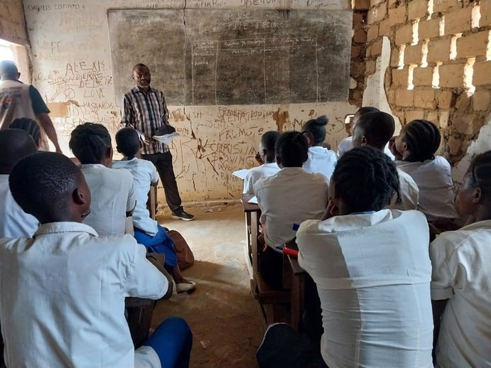 A teacher stands at the front of a classroom with brick walls and a chalkboard, instructing students seated at wooden desks, all wearing white uniforms.